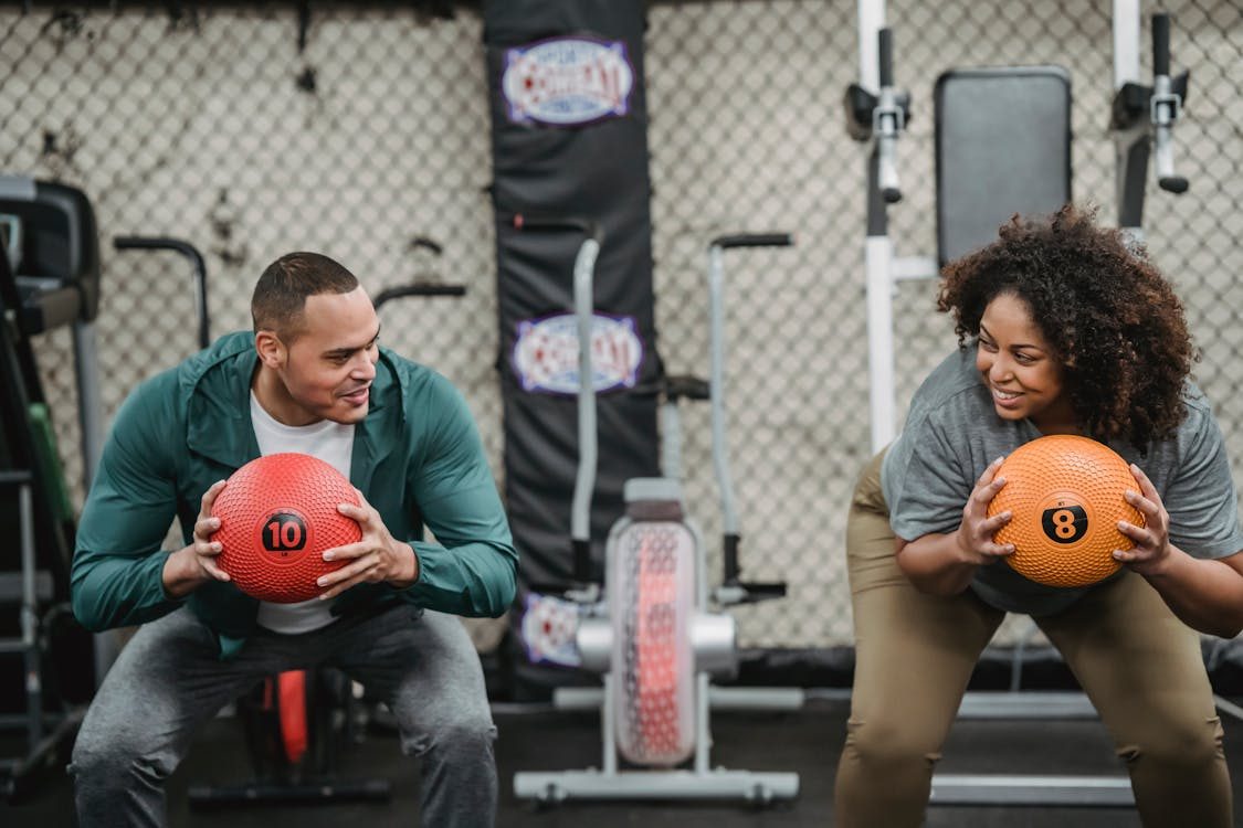 Two people squatting and smiling while holding weighted medicine balls in a gym, surrounded by exercise equipment and posing as if sharing a fun workout.