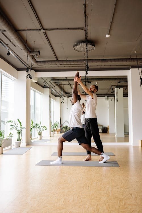 A yoga instructor helps a person in a lunge pose during a yoga session in a bright studio with large windows and potted plants.