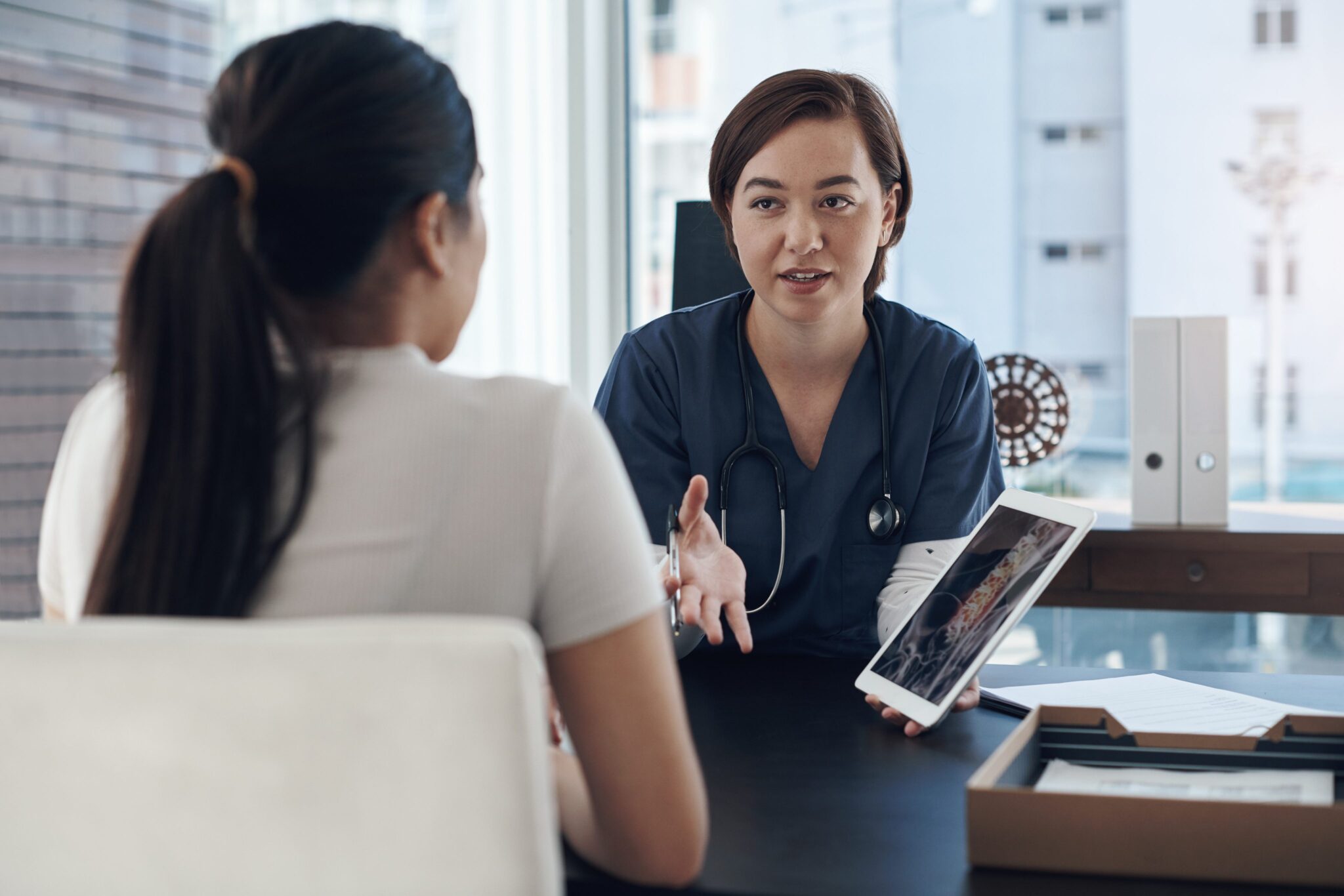 A healthcare professional in scrubs discusses medical information on a digital tablet with a patient across a desk in a bright office.