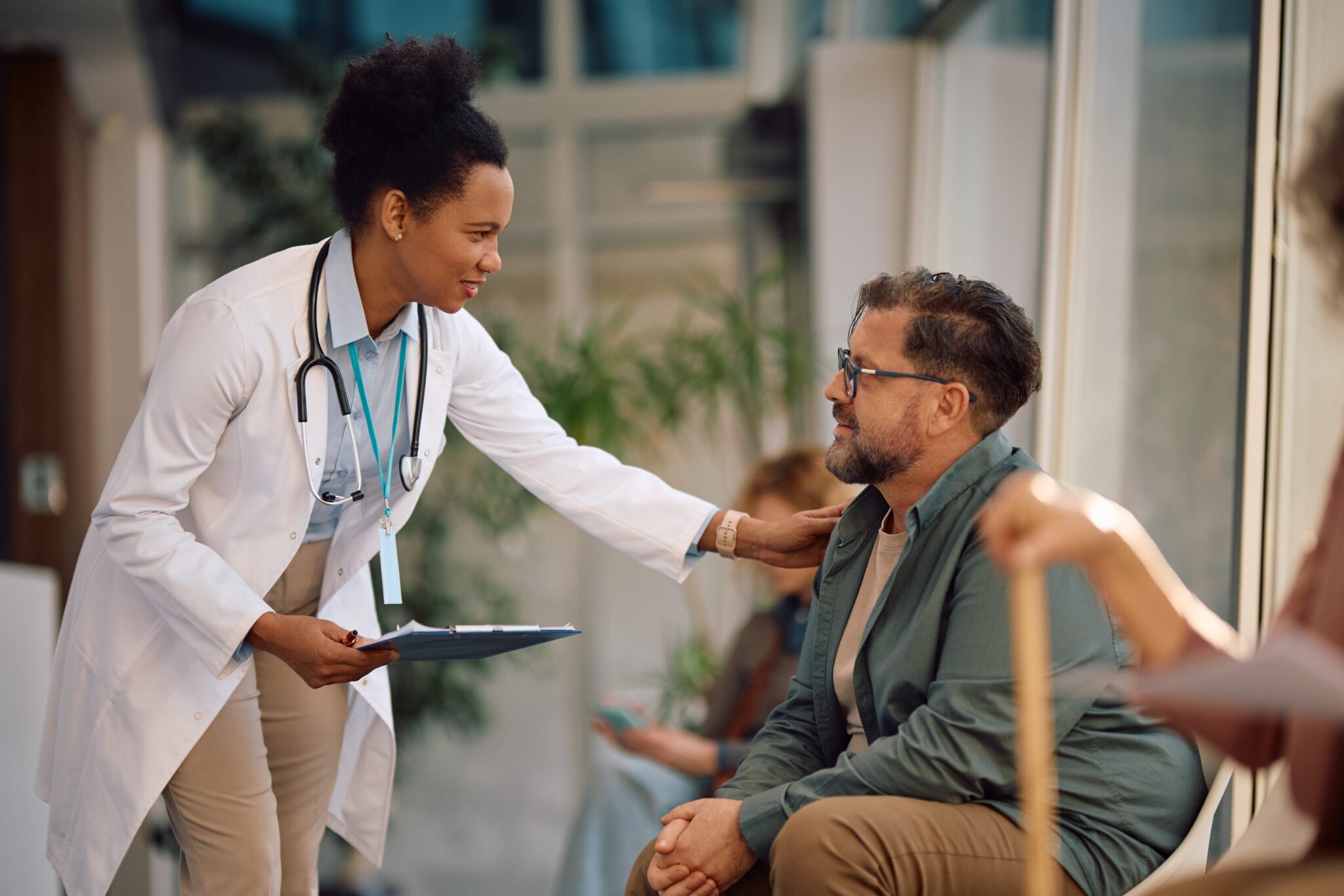 A doctor with a stethoscope and clipboard gently touches a seated man's shoulder, offering comfort. They are in a bright medical waiting area with plants and large windows.