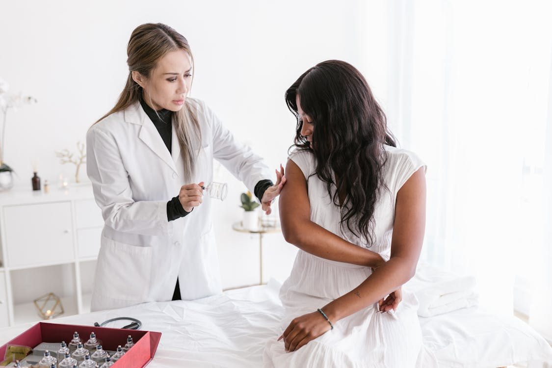 A female healthcare professional in a white coat prepares to administer an injection to a woman sitting on a medical bed in a bright, clean clinic setting.