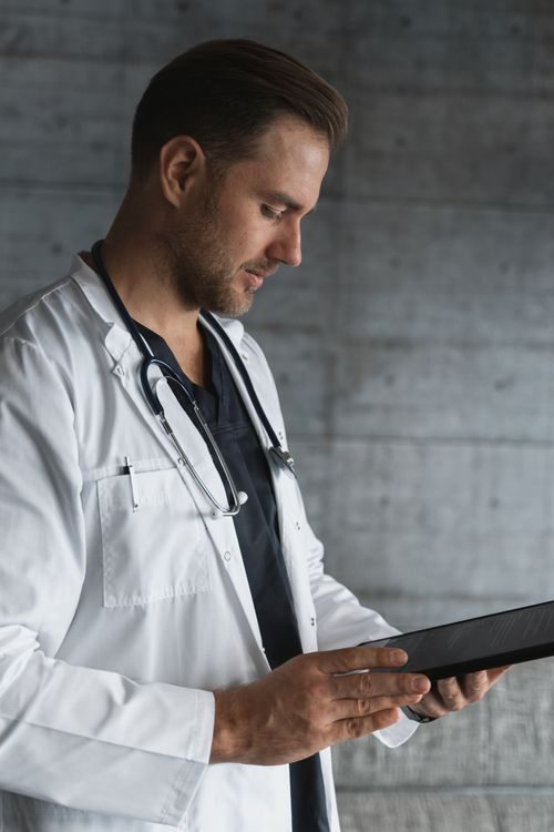 A male doctor wearing a white coat and stethoscope looks at a digital tablet, appearing focused, with a grey concrete wall in the background.