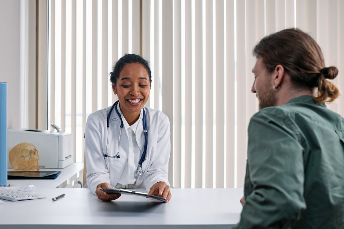 A smiling doctor in a white coat and stethoscope is sitting at a desk, holding a digital tablet and talking to a patient across from her in a medical office with vertical blinds in the background.