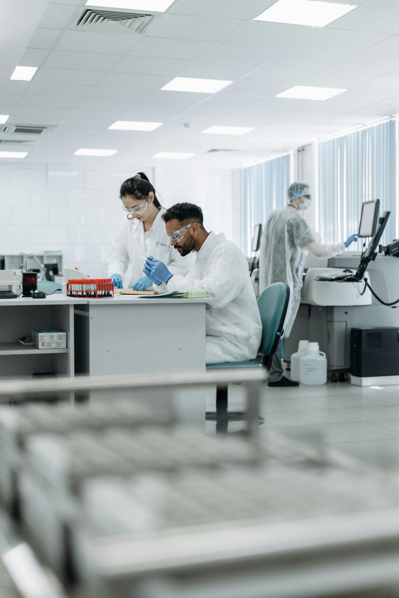 Two scientists in protective gear work at a lab desk with equipment and samples, while another person uses a computer in the background in a bright, modern laboratory.