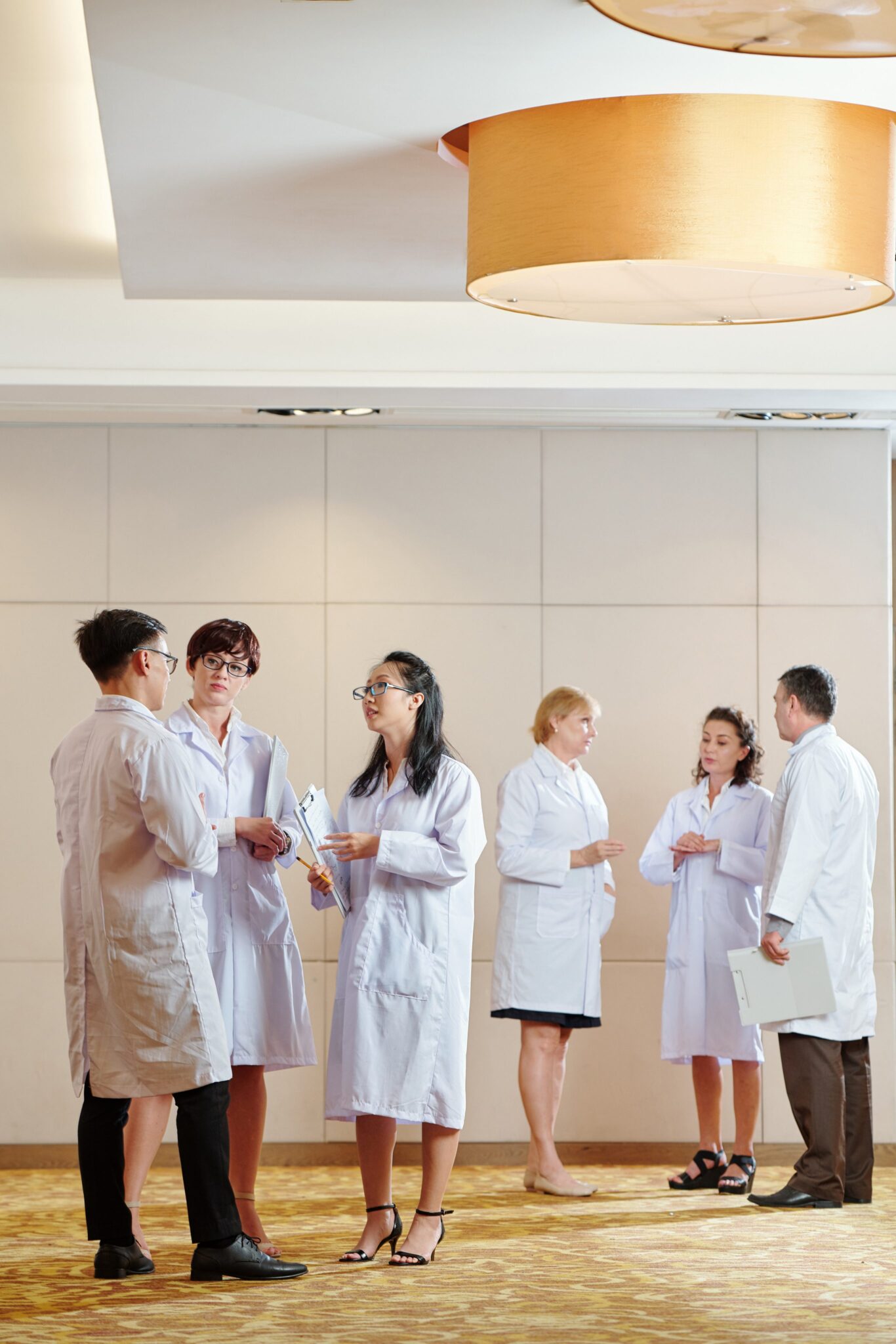 A group of six people in white lab coats stand in pairs, talking and holding clipboards in a bright, modern room with beige walls and carpet.