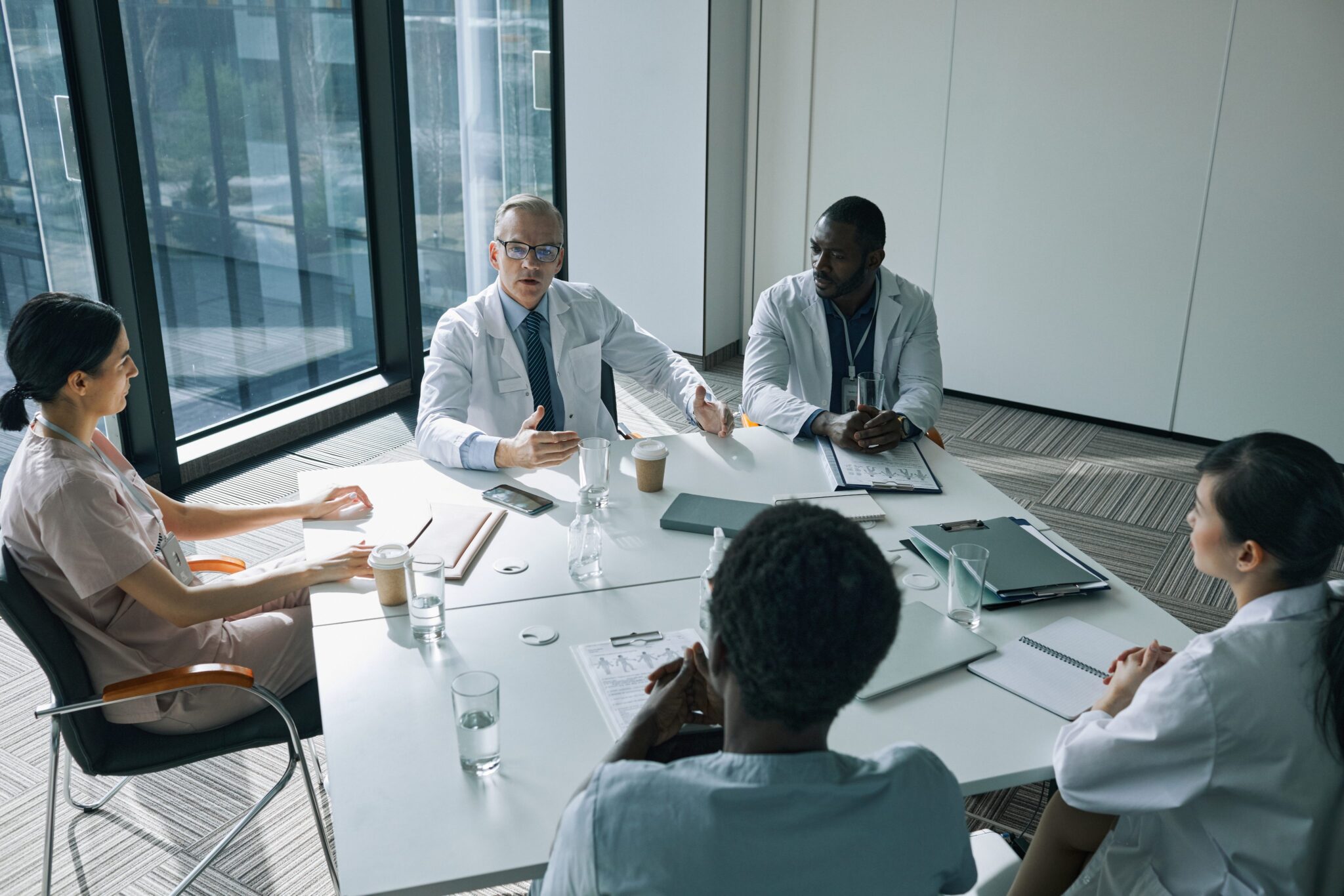 Five medical professionals sit around a conference table in a modern office, engaged in discussion. Papers, laptops, and drinks are on the table. Large windows provide natural light.