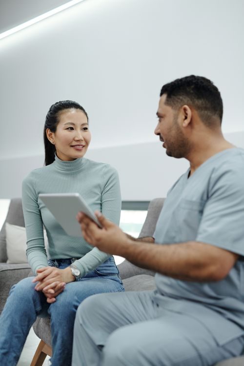 A woman sits and smiles while listening to a healthcare professional in scrubs, who is holding a digital tablet and speaking to her in a modern office setting.