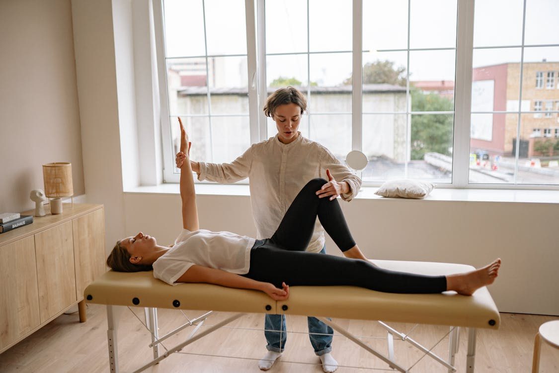 A physical therapist guides a woman lying on an examination table through a stretching or mobility exercise, lifting her arm and leg in a bright room with large windows.
