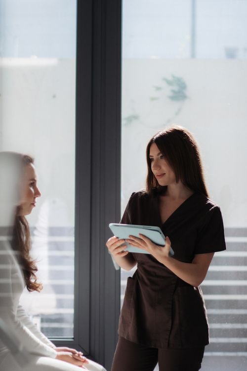 A woman in medical scrubs holds a tablet and talks to another woman seated by a large window in a sunlit room.