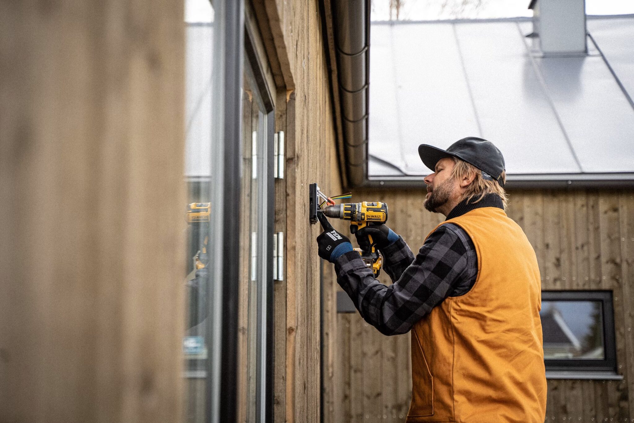 A man wearing a black cap, orange vest, and plaid shirt uses a power drill to install something on the exterior wooden wall of a building with large windows.