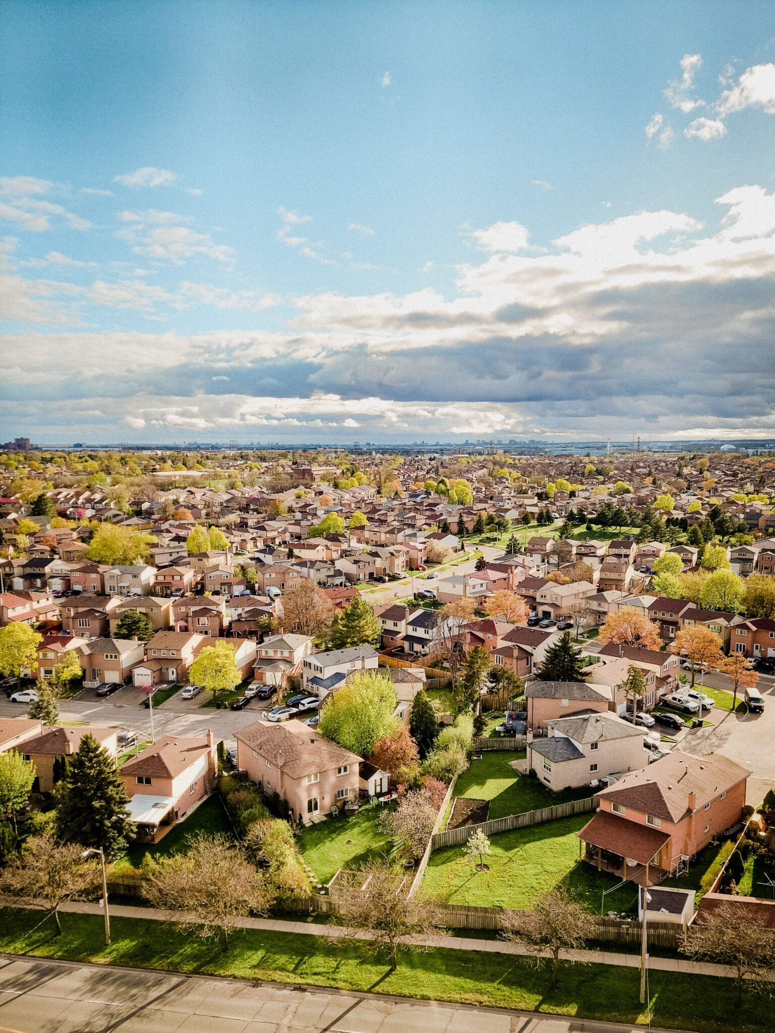Aerial view of a suburban neighborhood with rows of houses, tree-lined streets, and green lawns under a partly cloudy sky on a bright day.