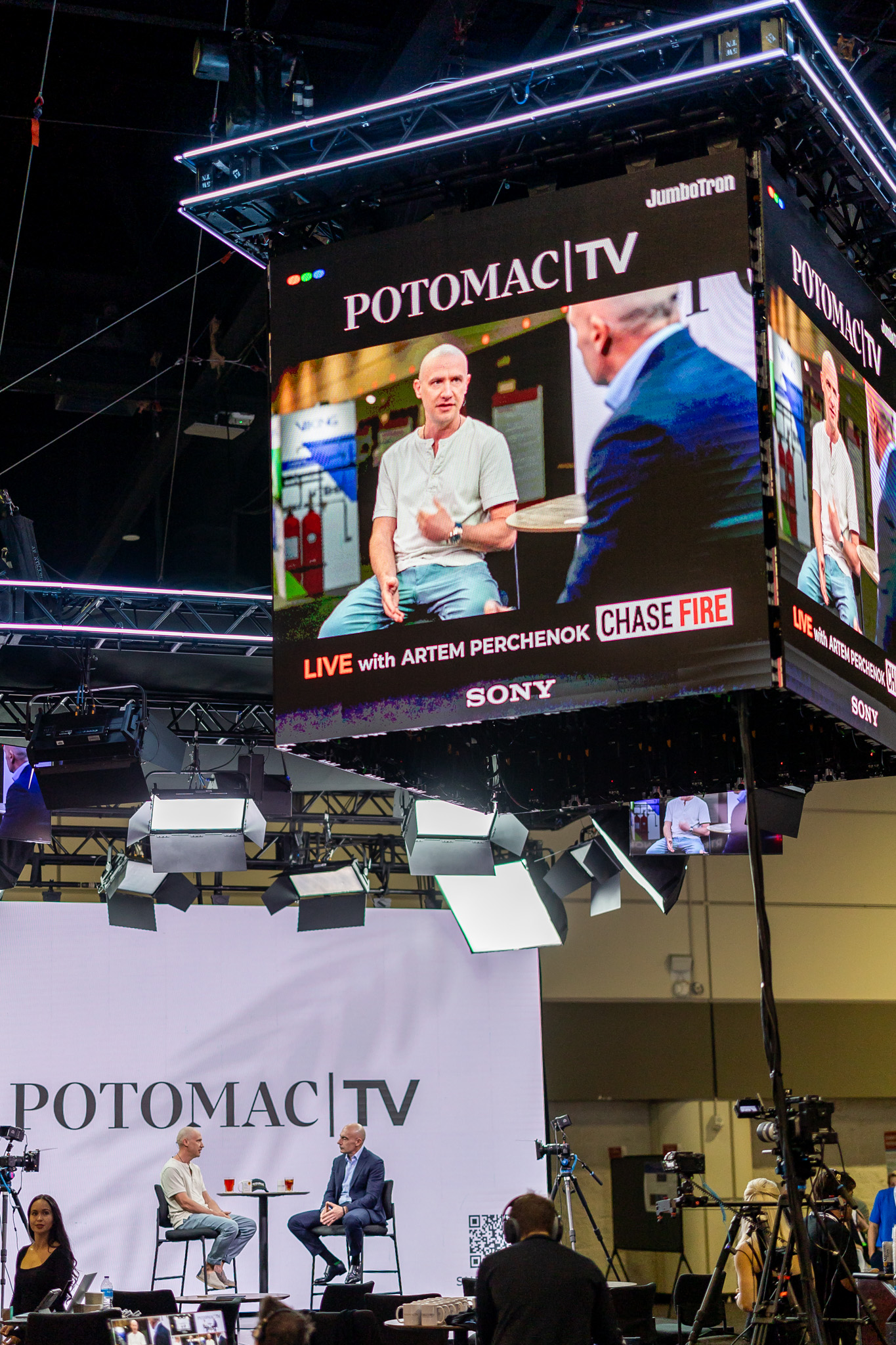 Two people sit and talk on a stage for POTOMAC TV while being filmed, with their discussion displayed on a large overhead screen. Studio lights and cameras surround the set.