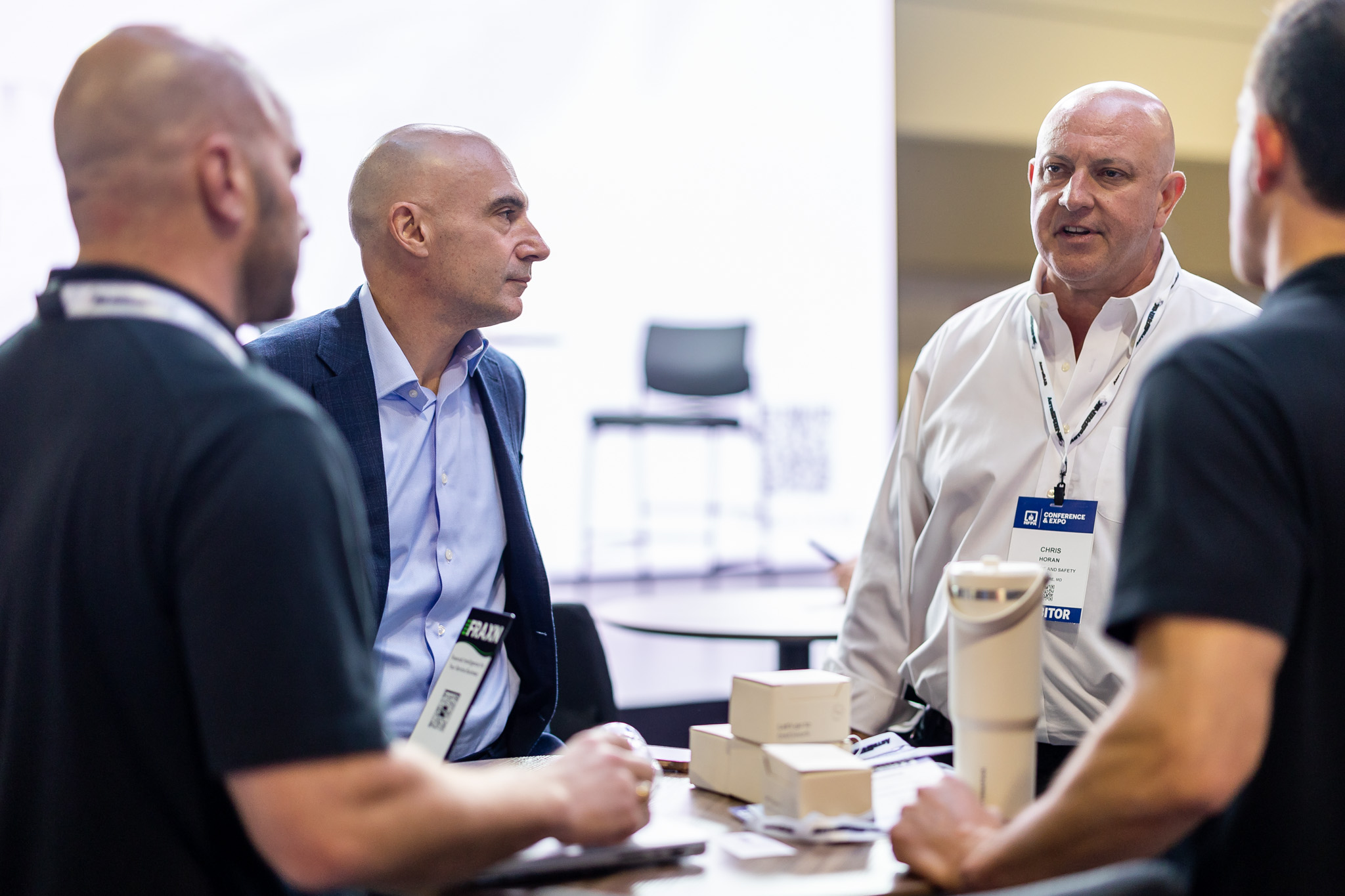 Four men in business attire stand around a table with boxes and a white device, engaged in conversation at a professional event. Two have visible name badges, and the background features a blurred chair and bright lighting.