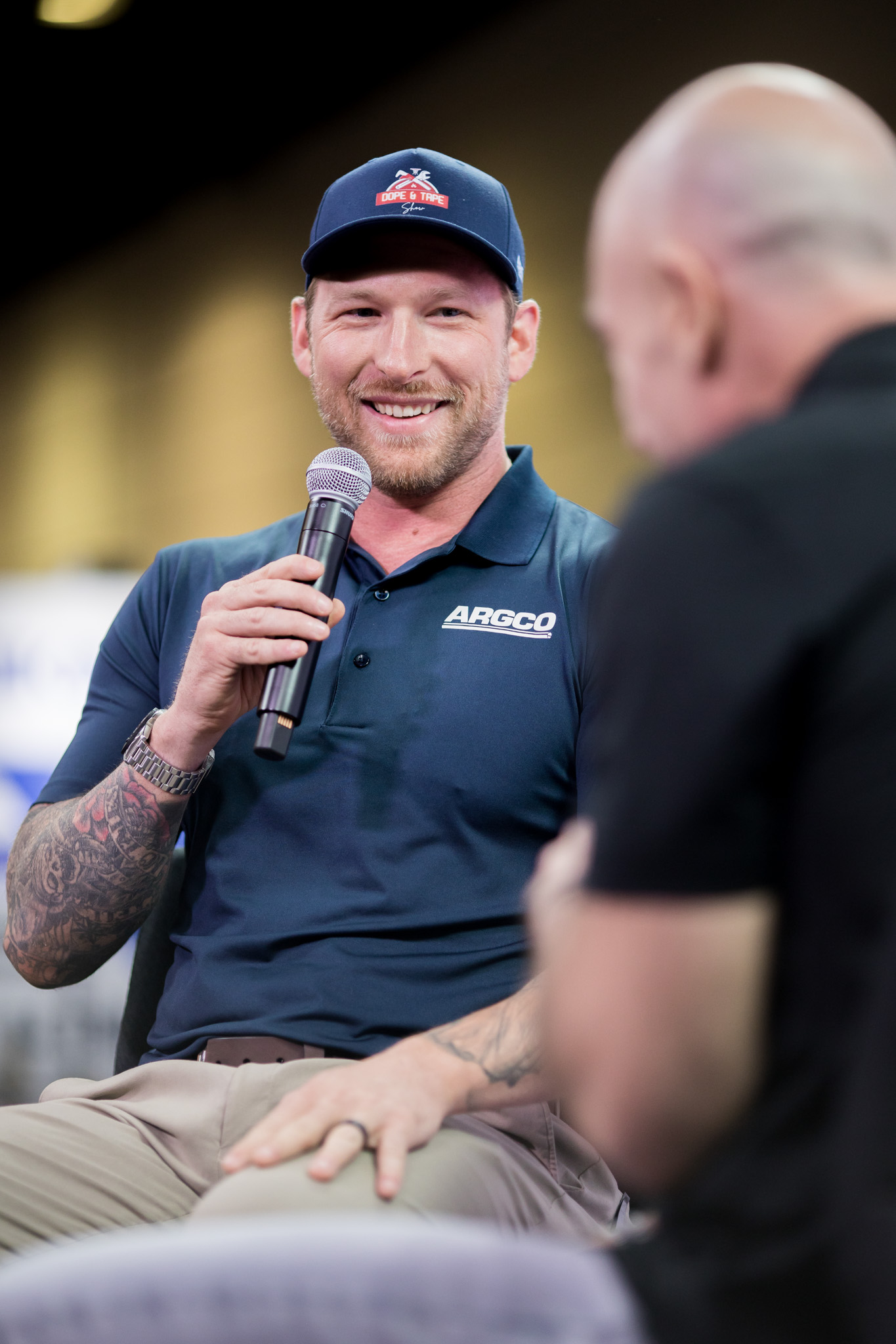 A smiling man with tattoos, wearing a navy polo shirt and cap, holds a microphone and speaks during an interview. Another person, blurred in the foreground, listens attentively.