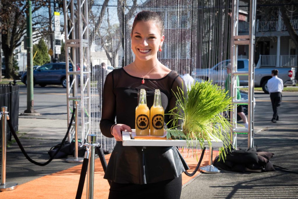 A smiling woman in black holds a tray with two bottles of juice featuring paw print labels and a bundle of grass. She stands on an orange carpet outdoors, with event decorations and people visible in the background.