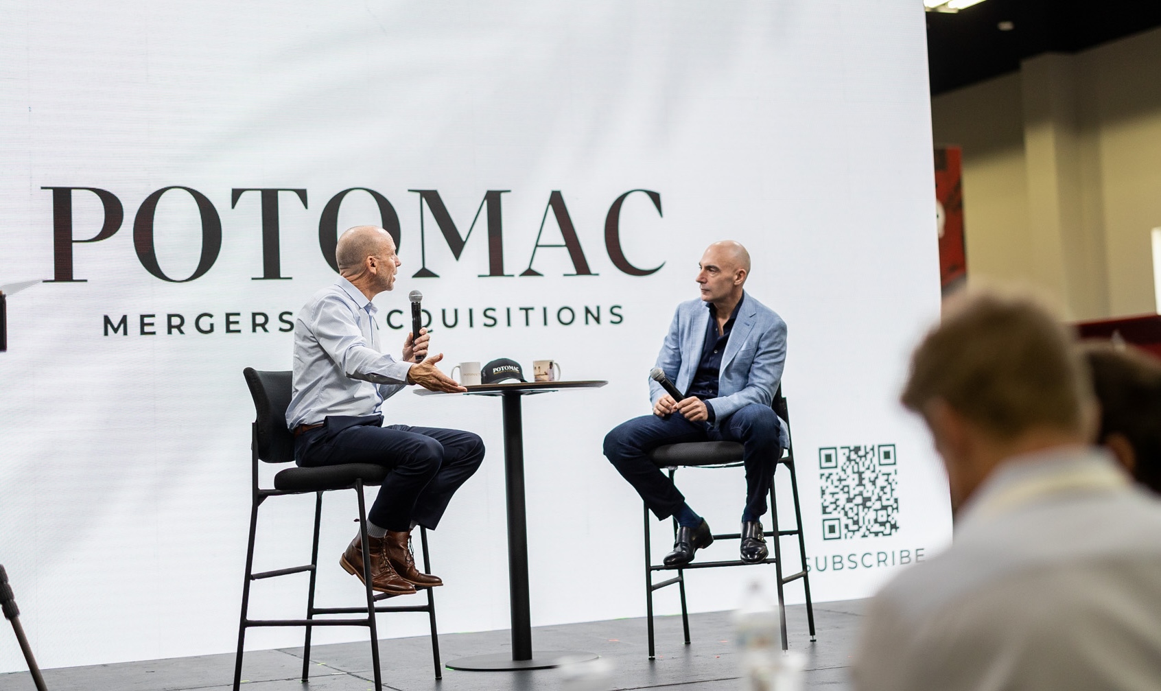 Two men sit on high chairs on a stage, engaged in conversation, with a large screen behind them displaying "Potomac Mergers & Acquisitions" and a QR code labeled "Subscribe." An audience is visible in the foreground.
