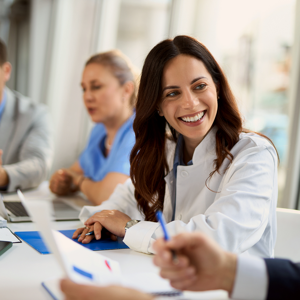 A smiling woman in a white lab coat sits at a table during a meeting, with other people in professional attire and scrubs in the background, suggesting a healthcare or medical setting.