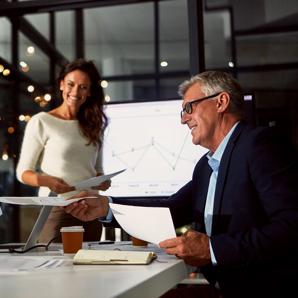 Two colleagues in a modern office share documents at a desk. A woman stands smiling, while a man in glasses sits and hands her papers. A graph is displayed on a screen behind them, and coffee cups are on the table.