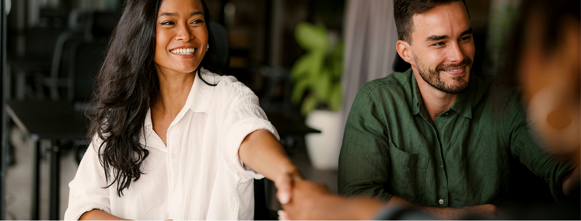 A smiling woman in a white shirt reaches out to shake hands with someone off-camera, while a man in a green shirt sits beside her, both appearing friendly and engaged in a professional setting.