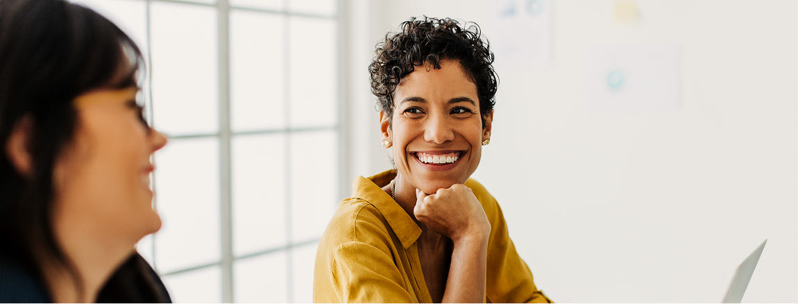 Two women are sitting indoors and talking. One woman with short curly hair and a yellow shirt is smiling brightly at the other woman. Natural light comes in through large windows in the background.