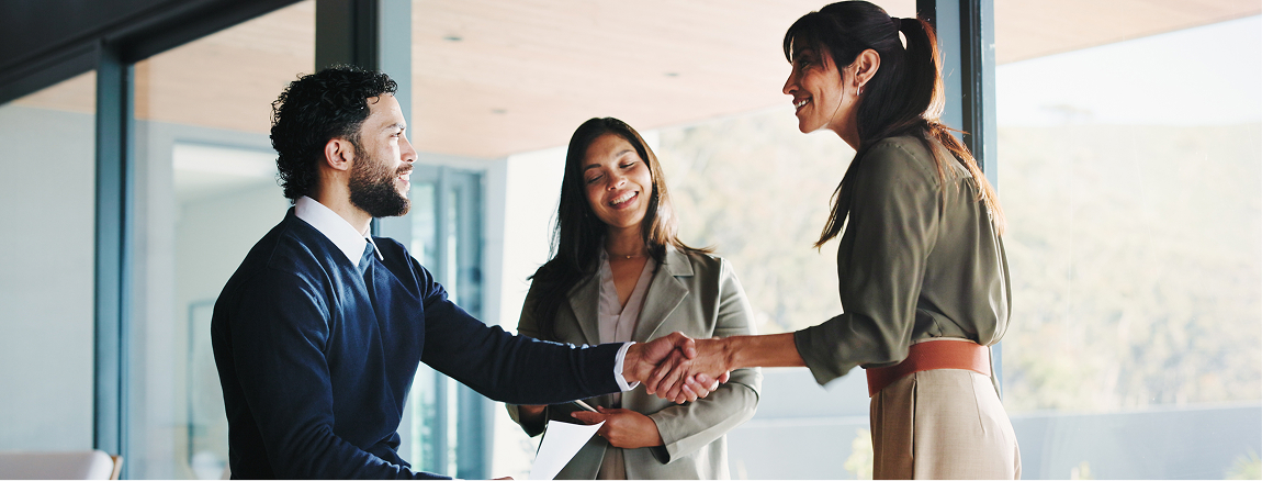 Three people in business attire are meeting in an office. Two are shaking hands and smiling, while the third stands behind them, also smiling, holding a clipboard or folder. Large windows reveal a bright, outdoor view.