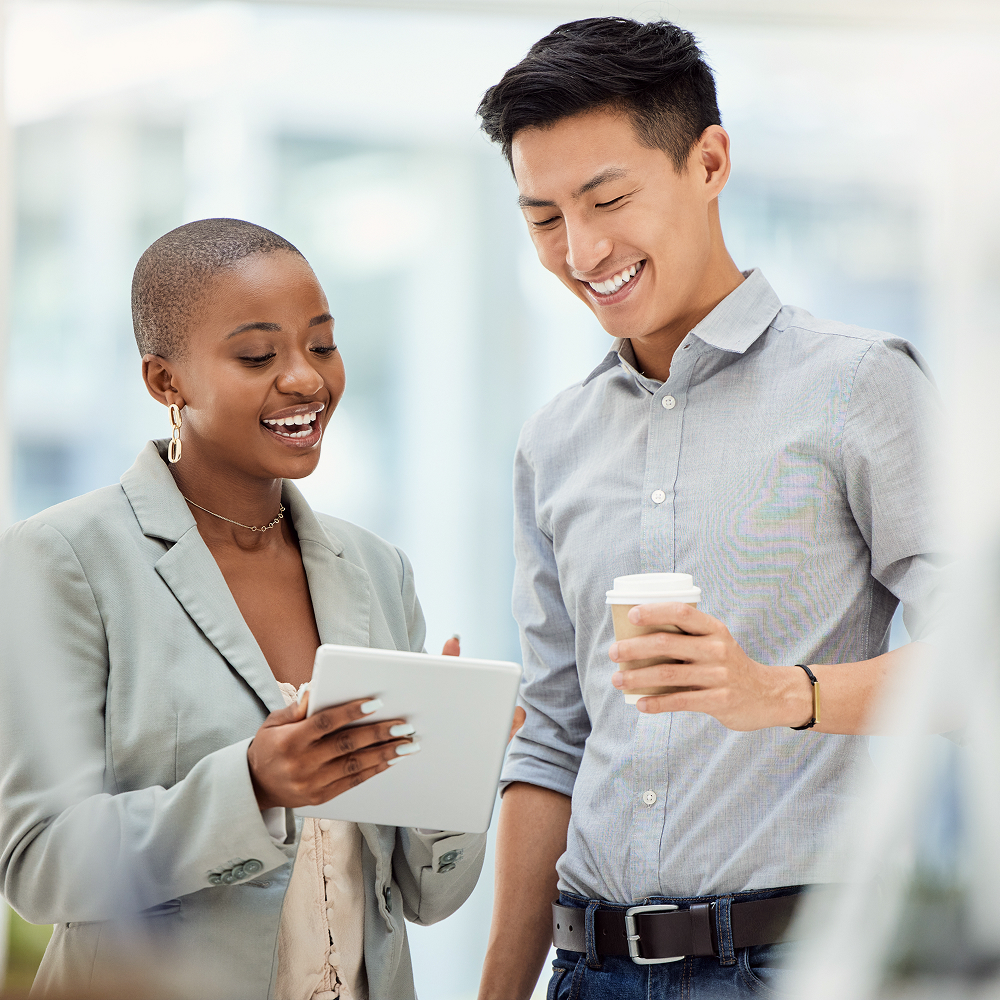 Two professionally dressed people smile while looking at a tablet. One holds the tablet and points at the screen, while the other holds a paper coffee cup. They appear to be having a positive conversation in a bright office.