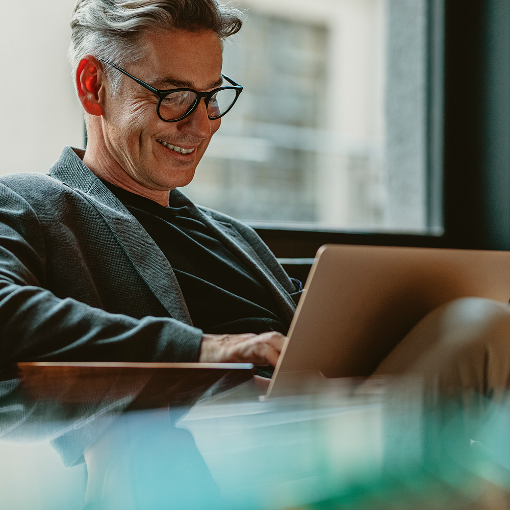 A smiling man with glasses and gray hair sits indoors, working on a laptop. He wears a dark blazer and black shirt, and natural light comes in through a large window behind him.