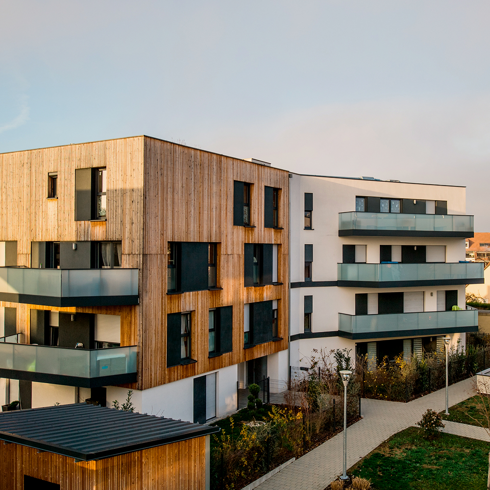 Modern apartment building with a mix of wooden and white exterior, large windows, and glass balconies. Pathways and landscaped greenery surround the structure under a partly cloudy sky.