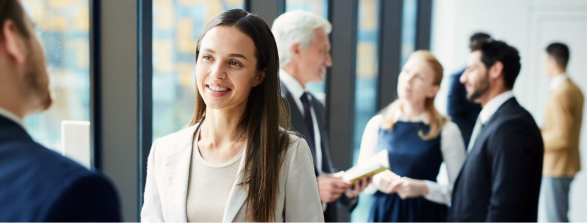 A woman in business attire smiles while talking to a colleague at a professional networking event. Other professionals are seen conversing in the background near large windows.