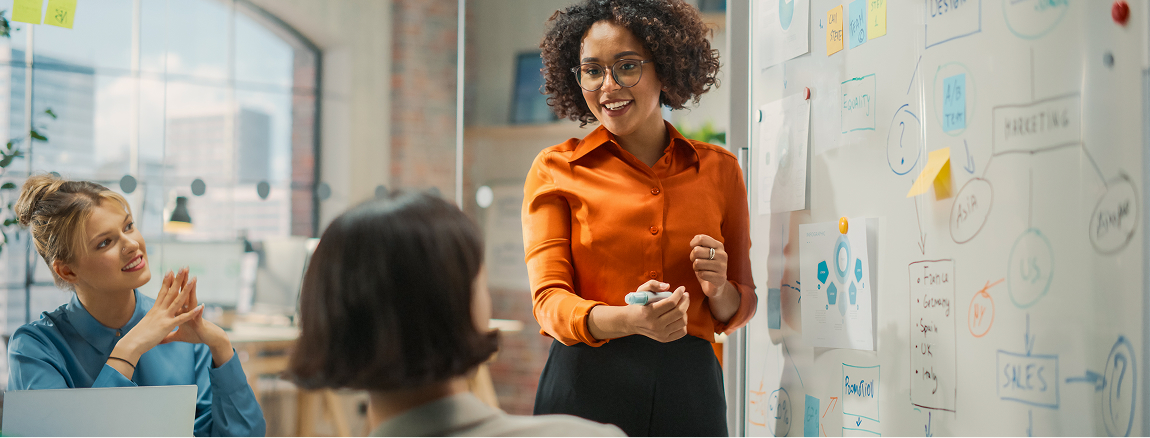 A woman in an orange blouse stands by a whiteboard covered with notes and diagrams, presenting to two colleagues seated and listening attentively in a modern office setting.