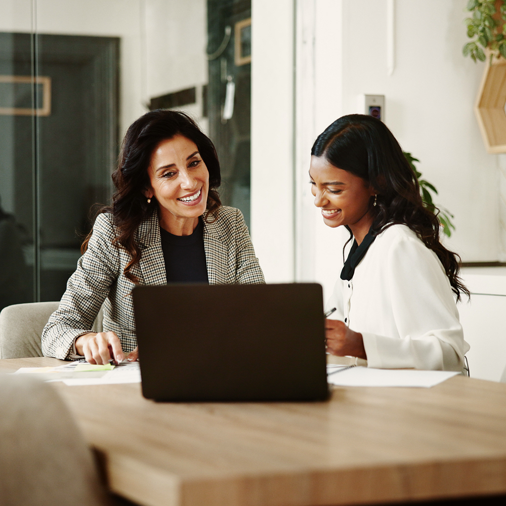 Two women sit at a desk smiling and looking at documents together, with an open laptop in front of them in a modern office setting.