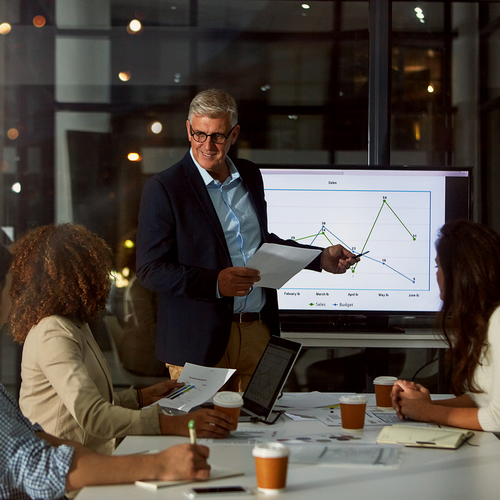 A man in a suit presents a line graph to three colleagues during a nighttime meeting in a modern office. The group is seated at a table with laptops, papers, and coffee cups.