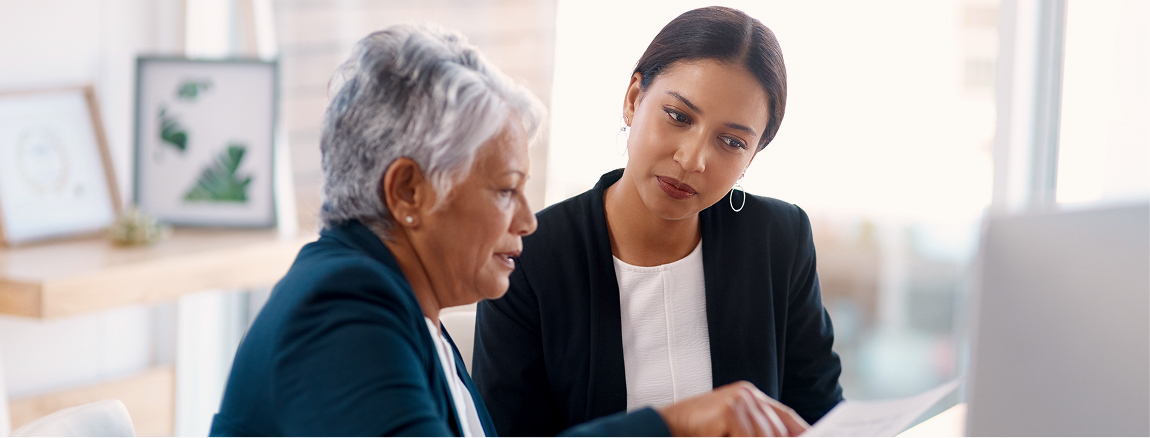 Two women in business attire sit at a desk, looking at documents together. One woman is older with short gray hair, the other is younger with dark hair tied back. They appear to be having a serious discussion in an office setting.
