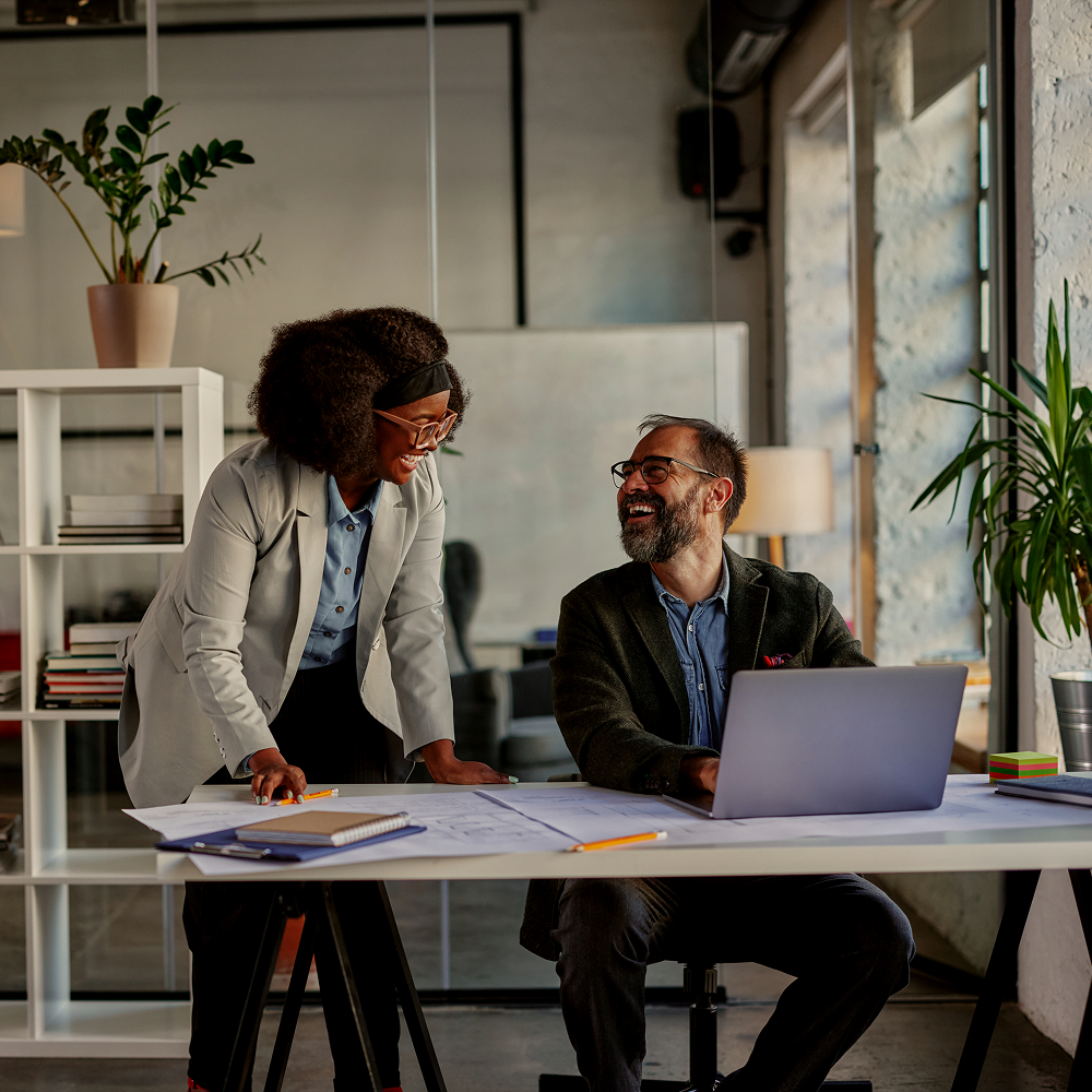 Two colleagues, a woman standing and a man sitting at a desk with a laptop, smile and talk in a modern office with large windows, plants, and shelves in the background.