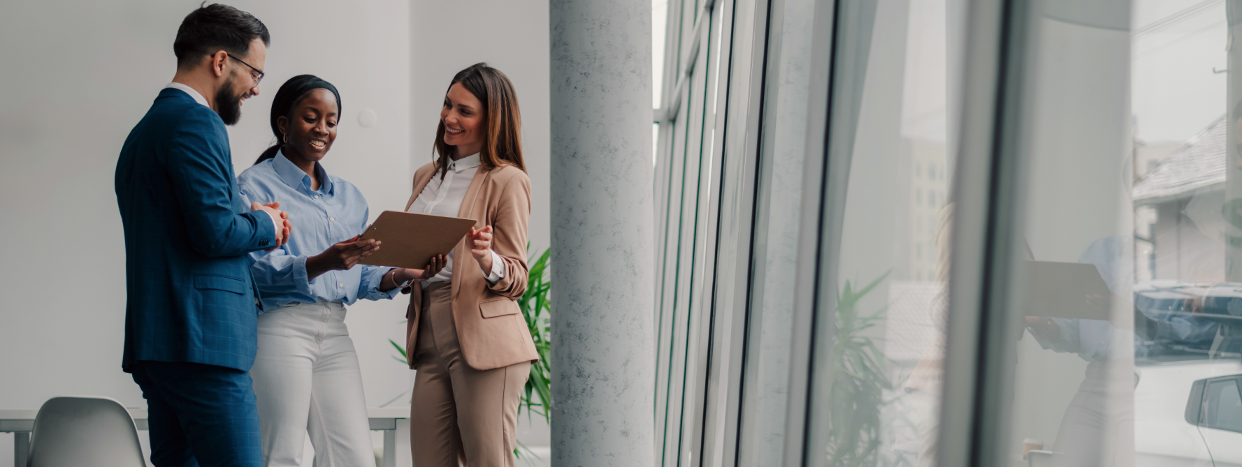 Three people in business attire stand together in a modern office by large windows, smiling and looking at a clipboard held by one of them.