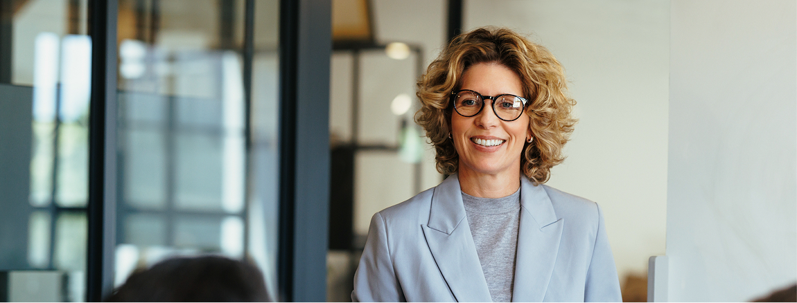 A smiling woman with curly blonde hair and glasses wearing a light gray blazer stands indoors in a modern office setting.