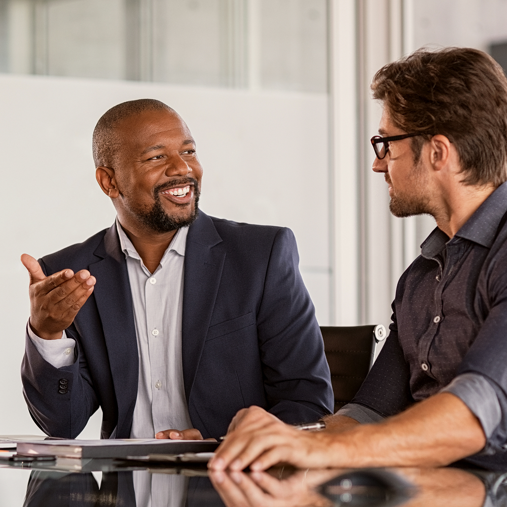 Two men sitting at a desk in an office, engaged in a friendly conversation. One man is smiling and gesturing with his hand, while the other listens attentively. Both are dressed in business attire.