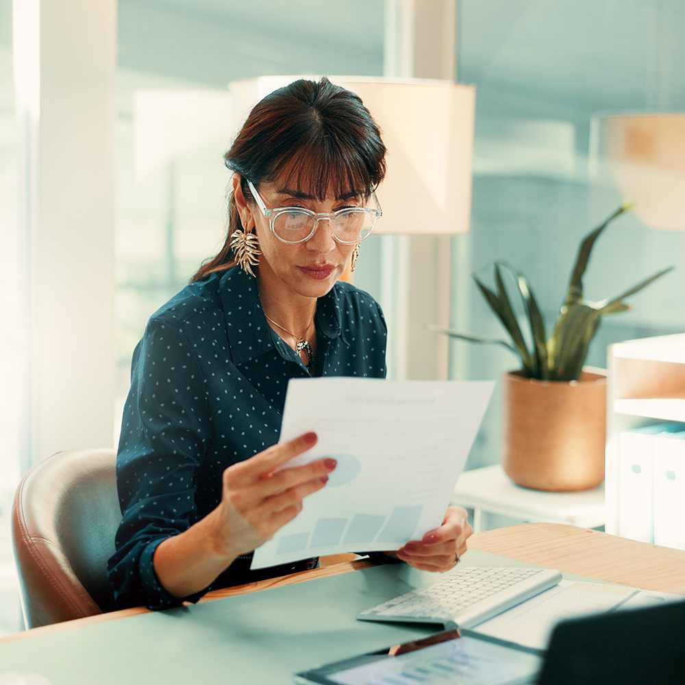 A woman wearing glasses and large earrings sits at a desk in a modern office, reviewing a document with graphs. A potted plant, lamp, and paperwork are visible in the background.