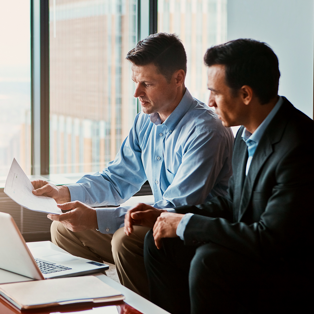 Two men in business attire sit by a window, looking at documents together. One man holds papers while the other listens attentively. A laptop is open on the table in front of them, with city buildings visible outside.