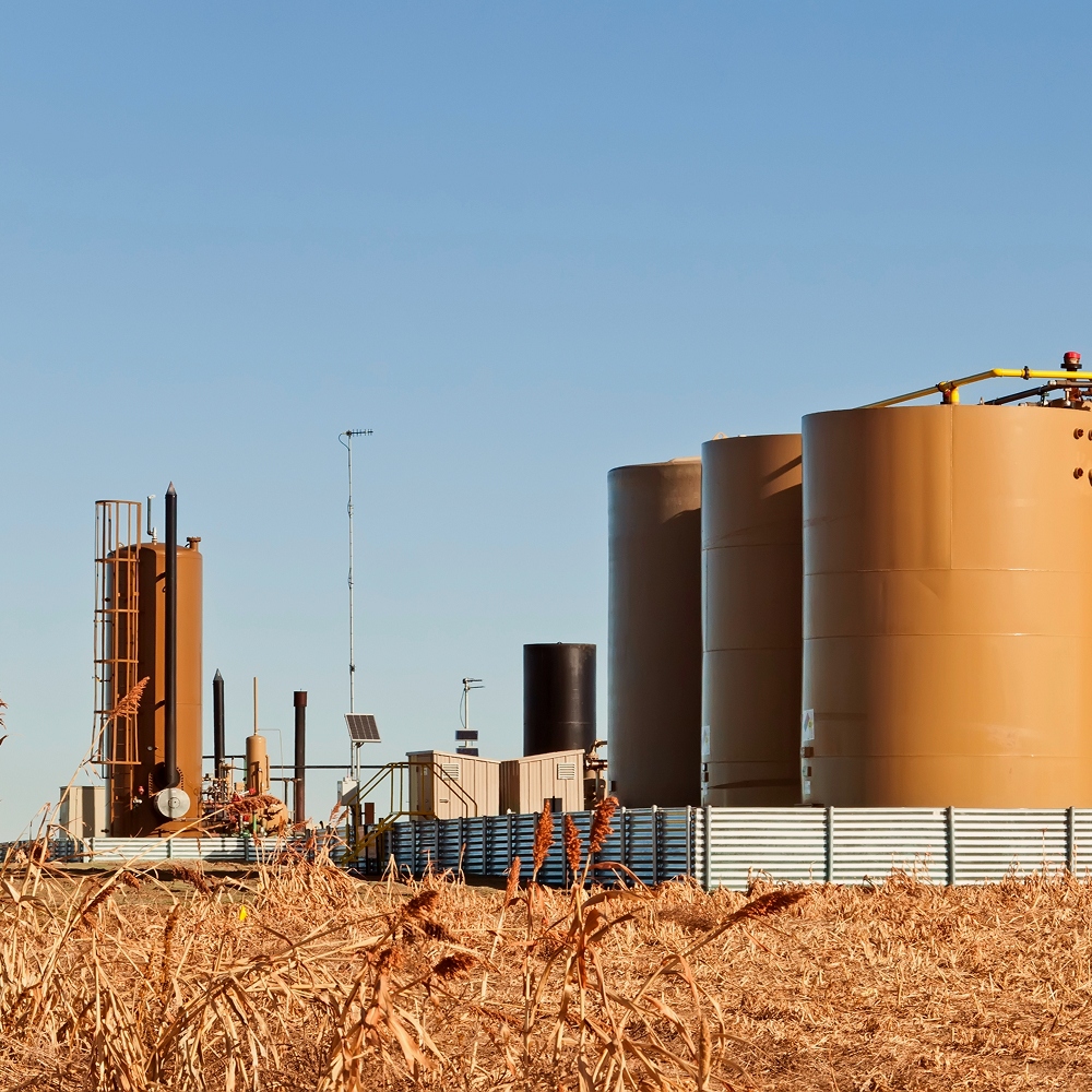 Several large, cylindrical storage tanks and industrial equipment are situated on a facility surrounded by dry, brown grass under a clear blue sky.