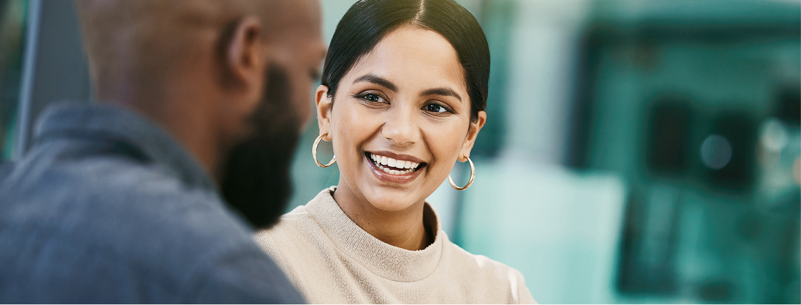 A woman with dark hair in a bun and gold hoop earrings smiles warmly at a man with a beard. They are having a conversation in a bright indoor setting with a blurred background.