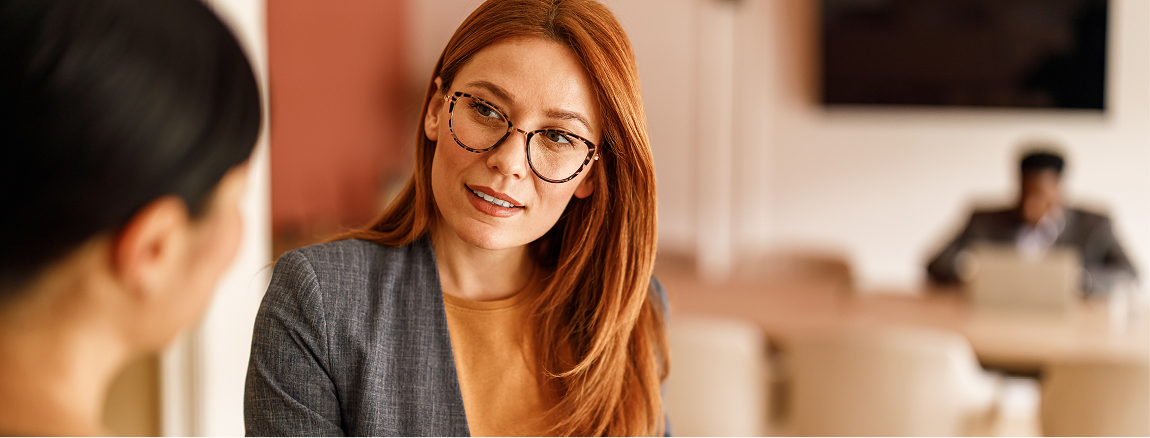 A woman with red hair and glasses, wearing a blazer, listens attentively to another person in an office setting. In the background, a person is working at a table with a laptop.