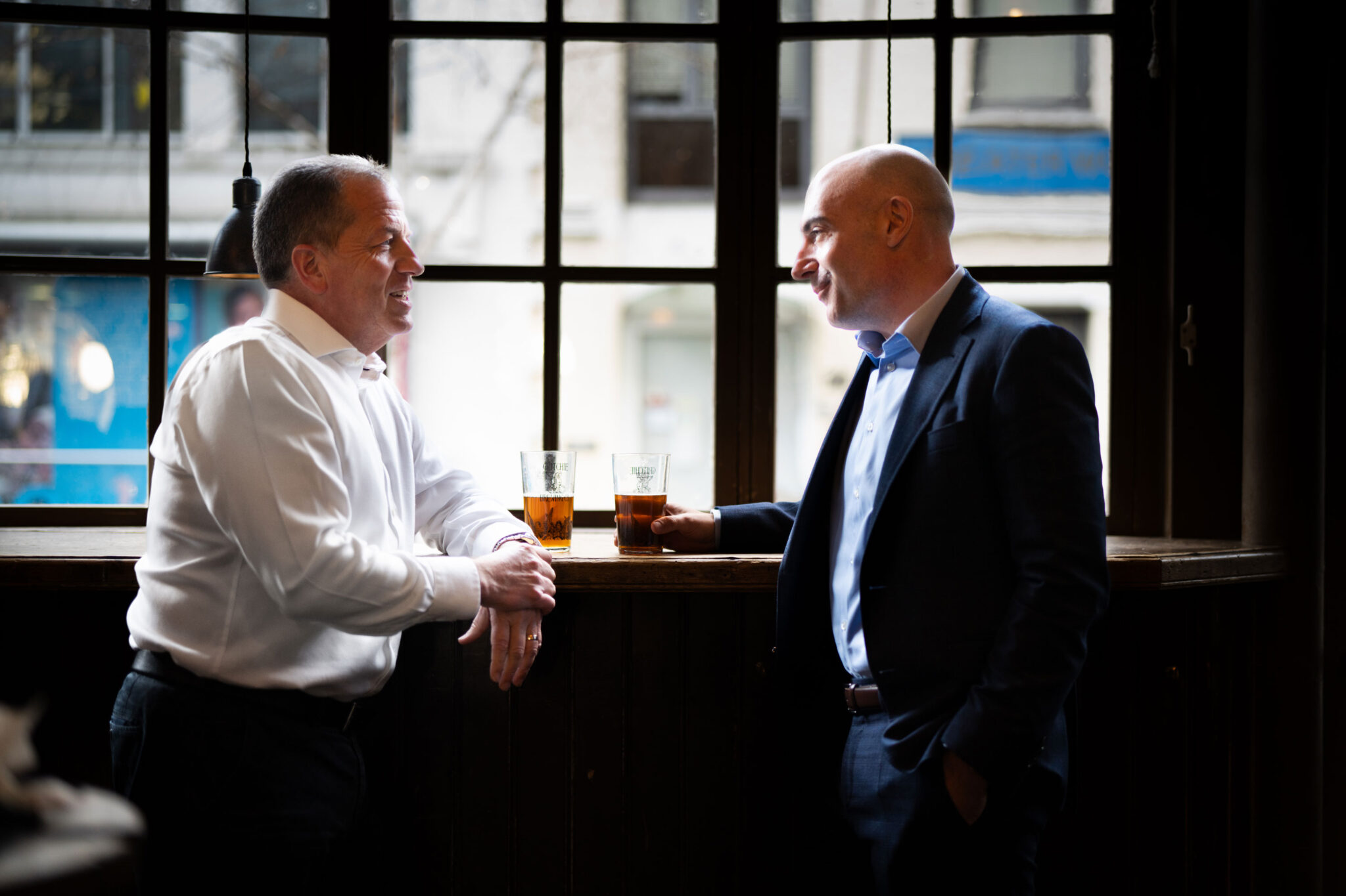 Two men in business attire stand by a window in a bar, each holding a glass of beer, engaged in conversation with natural light streaming in from outside.