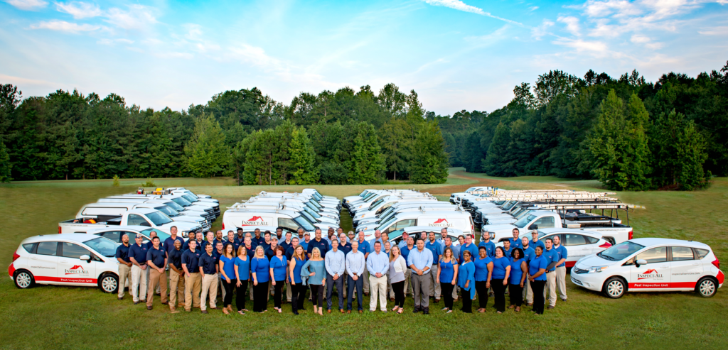 A large group of people stands in front of numerous white work vehicles parked on grass, with trees in the background and a blue sky above. The vehicles have company logos on their doors.
