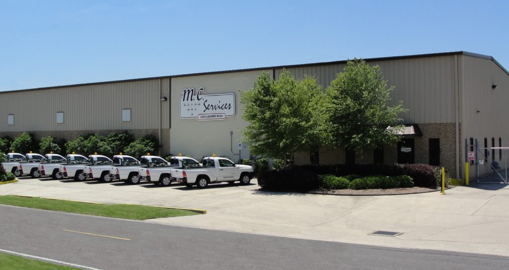A row of white pickup trucks is parked outside a large beige industrial building with a sign reading "MC Services." Bushes and trees border the front entrance under a clear blue sky.
