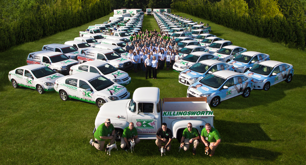A large group of people stands on grass, surrounded by many company-branded vehicles arranged in a V shape, with four people kneeling in front of a vintage company truck marked "Killingsworth.
