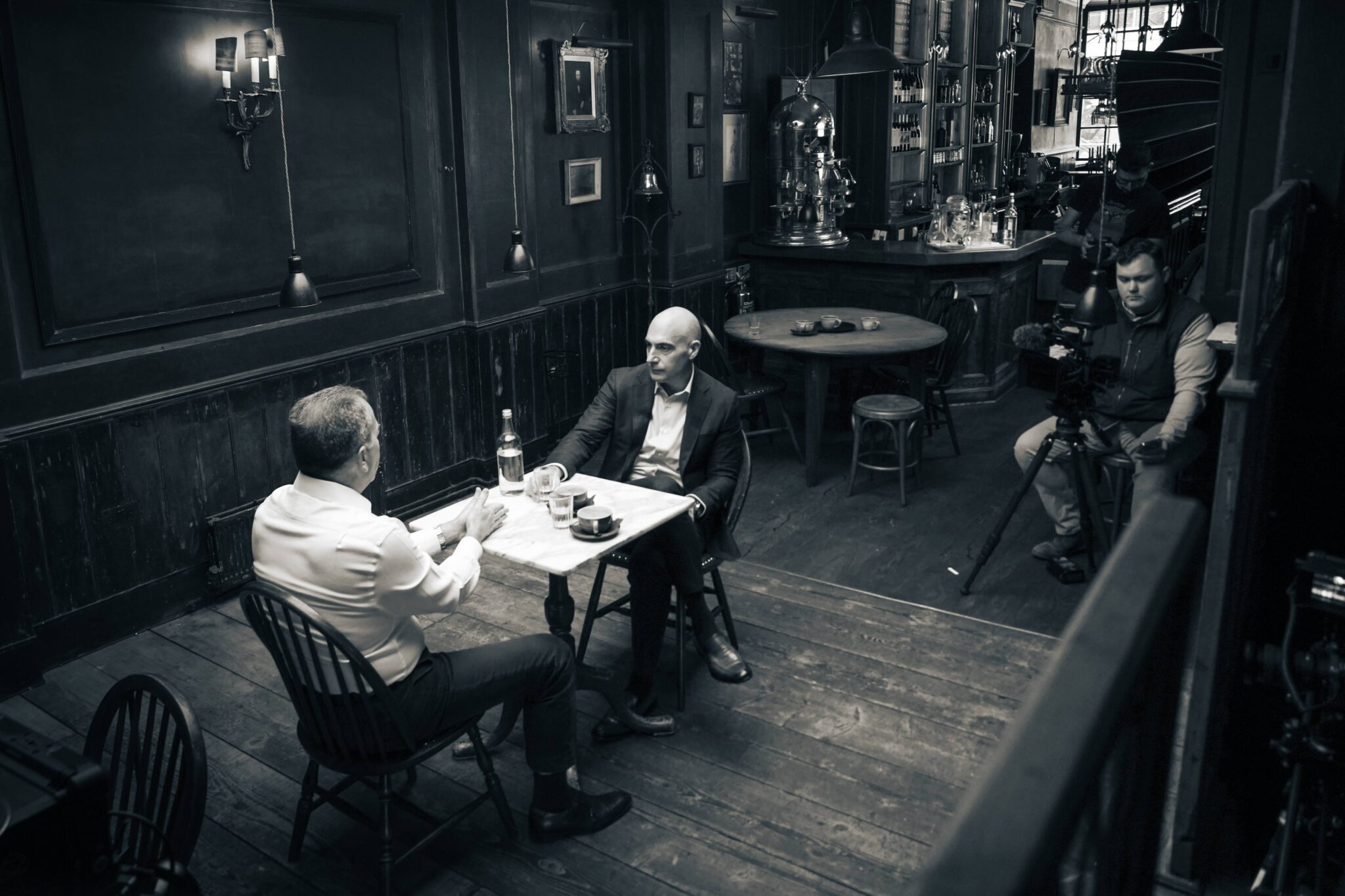 Two men sit across from each other at a small table in a dimly lit, vintage-style café while a cameraman films them. The scene appears to be an interview or conversation.