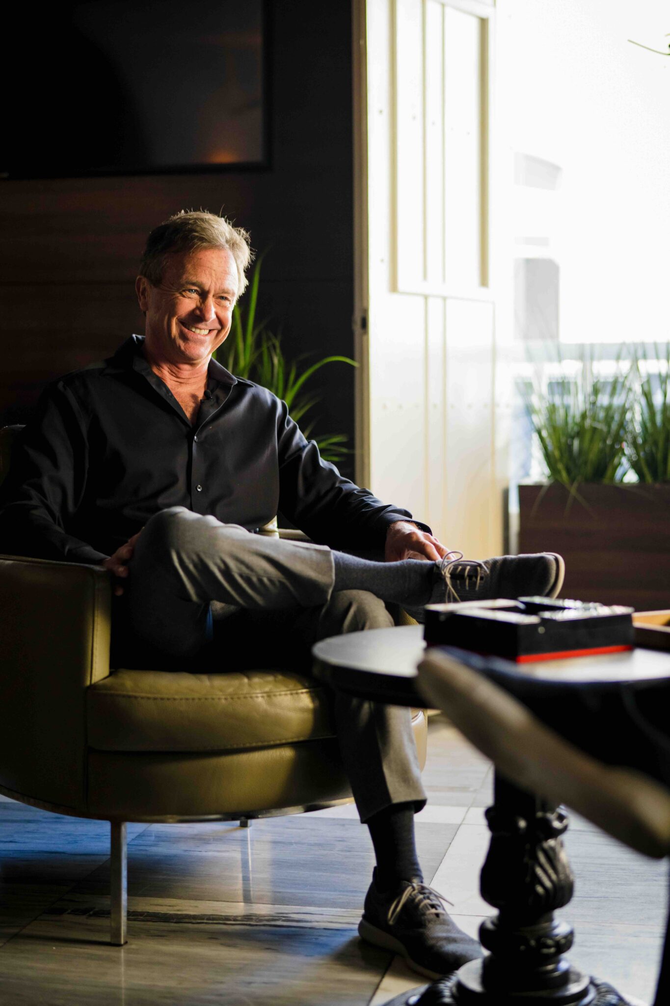 A man wearing a black shirt and gray pants sits relaxed in a chair with one leg crossed over the other, smiling, in a well-lit room with plants and a small table in the foreground.