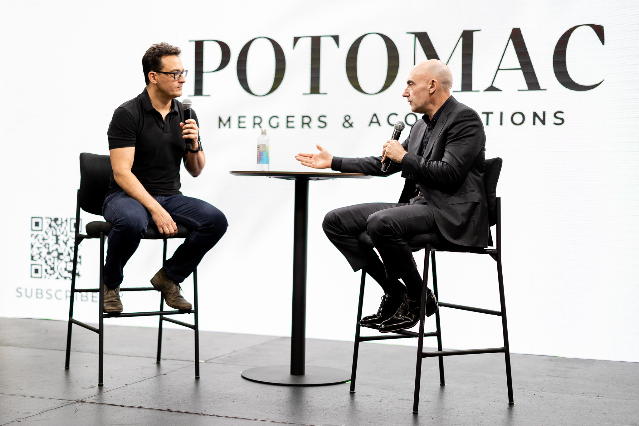Two men sit on tall chairs at a round table, having a discussion with microphones. Behind them is a large sign reading "POTOMAC MERGERS & ACQUISITIONS." A QR code is visible on the display.