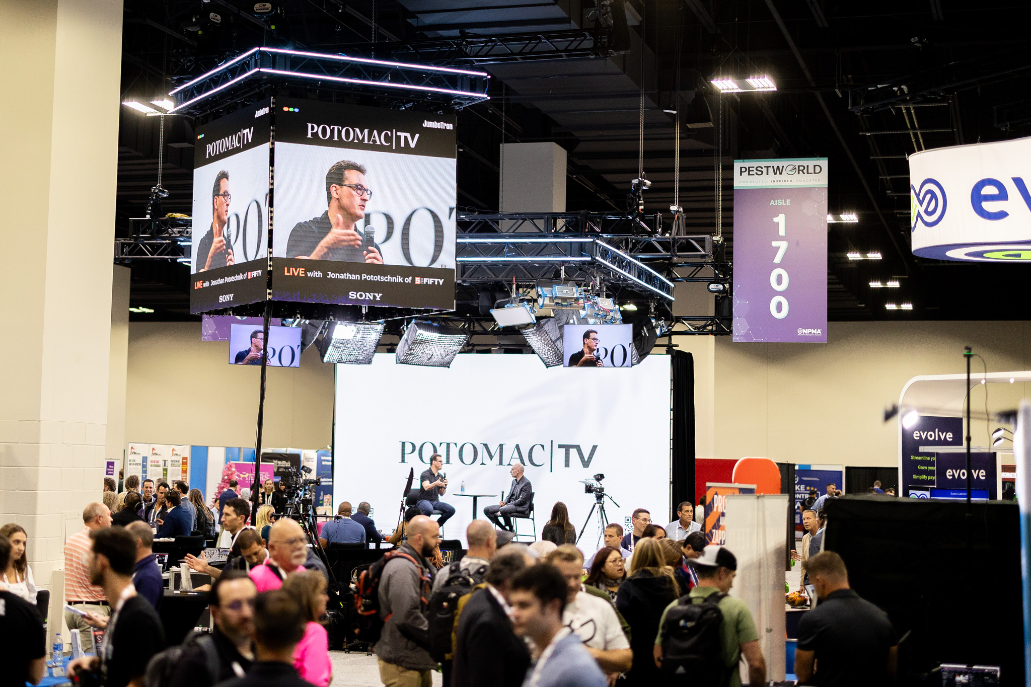 A busy convention hall with people walking around booths. A live interview is being conducted on a stage labeled "POTOMAC TV," displayed on a large screen above. Signs indicate the event and aisle number 1700.